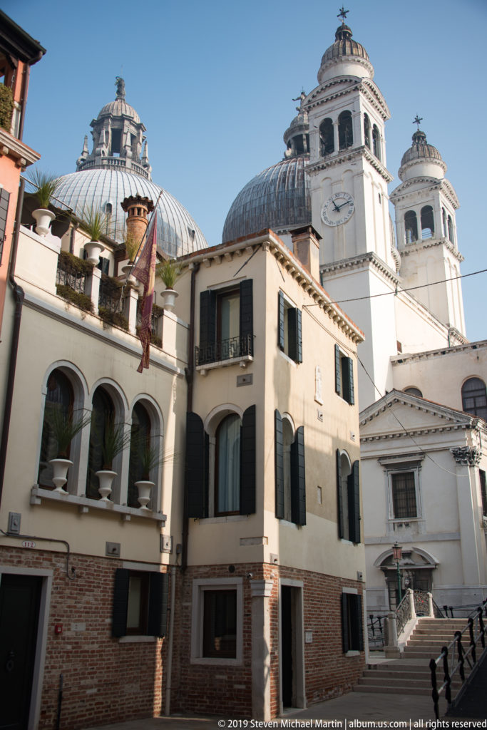 Large pale domed church rising behind brick and stucco Venetian houses with dark shutters.