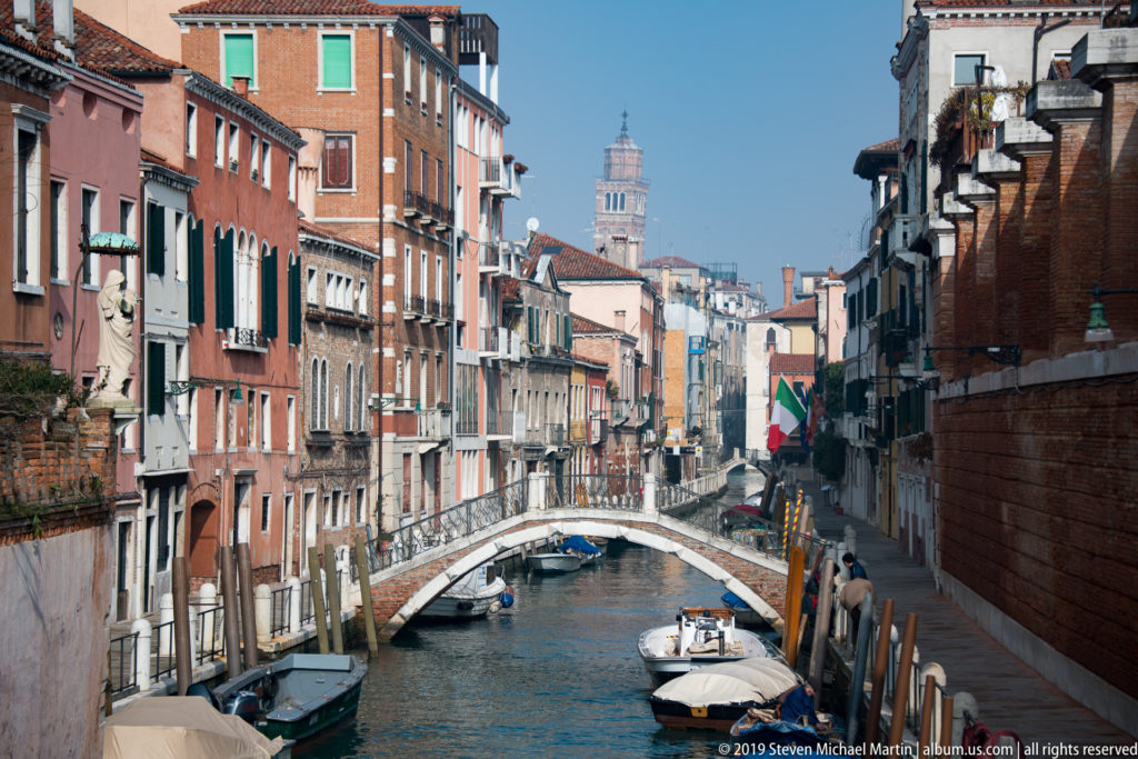 Arched pedestrian bridge over a Venetian canal lined with colorful buildings, mooring posts, boats, and a distant bell tower.