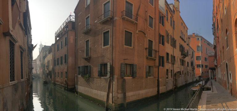 Narrow green canal running between tall stucco and brick buildings with shuttered windows in Venice.