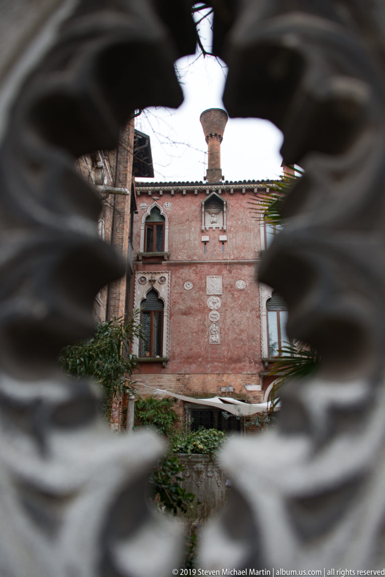 Red-brick Venetian residence and courtyard seen through blurred openings in a carved stone balustrade.