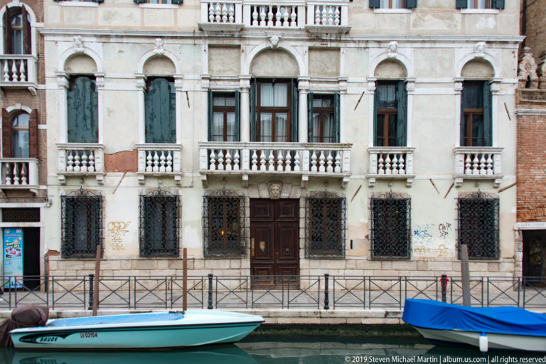 White canal-side palazzo with green shutters, small stone balconies, iron-grilled lower windows, central doors, and boats along the water.