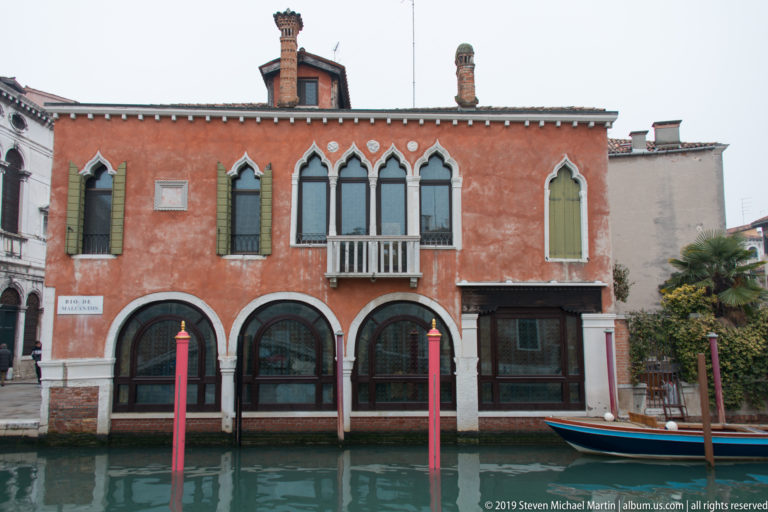 Red canal-side building with three broad lower arches, pointed upper windows, a small balcony, and mooring poles in front.