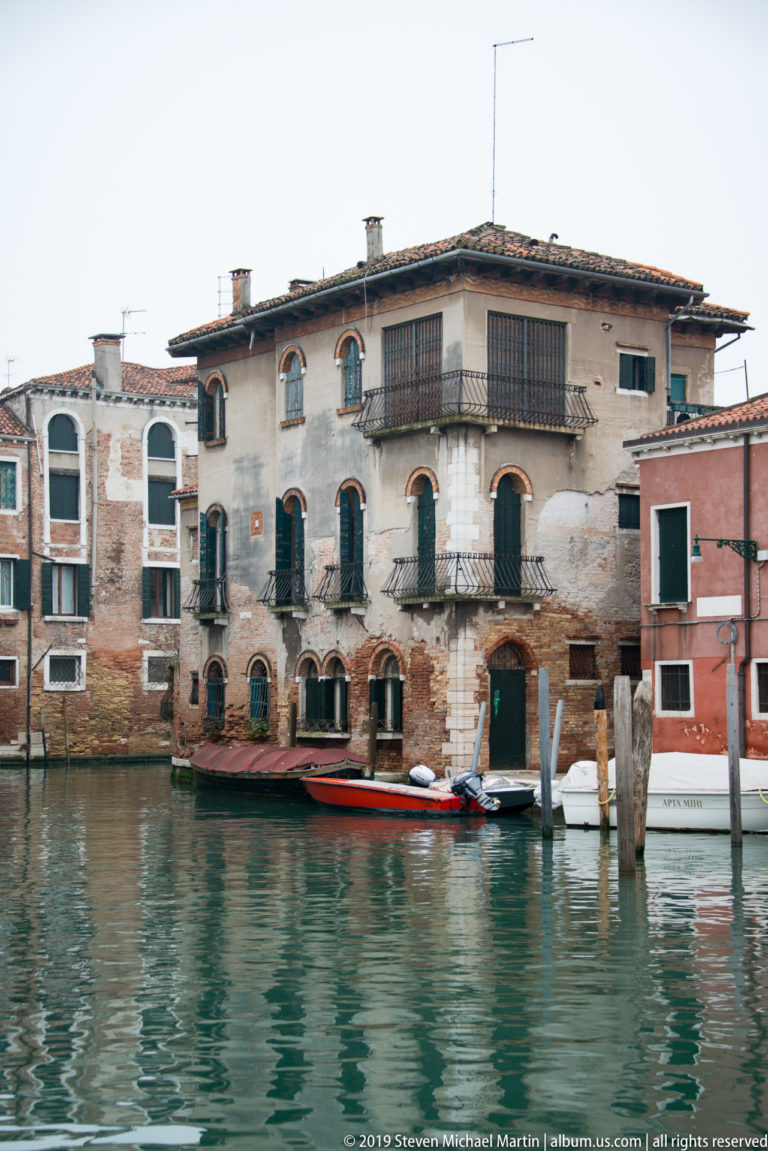 Weathered canal-side building with arched windows, balconies, green shutters, mooring posts, and small boats along the water.