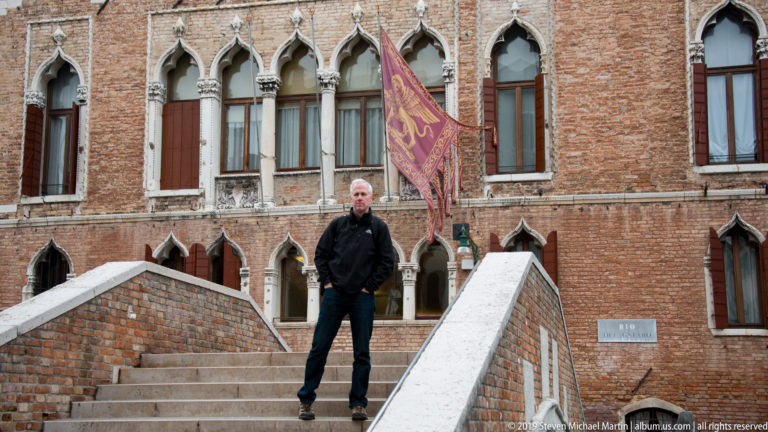 Man standing on a small brick footbridge before a red-brick Venetian Gothic building with pointed windows and a winged-lion banner.