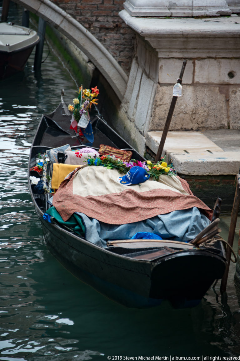 Low black workboat moored beside stone steps, covered with layered tarps and carrying flowers, bags, and assorted gear.