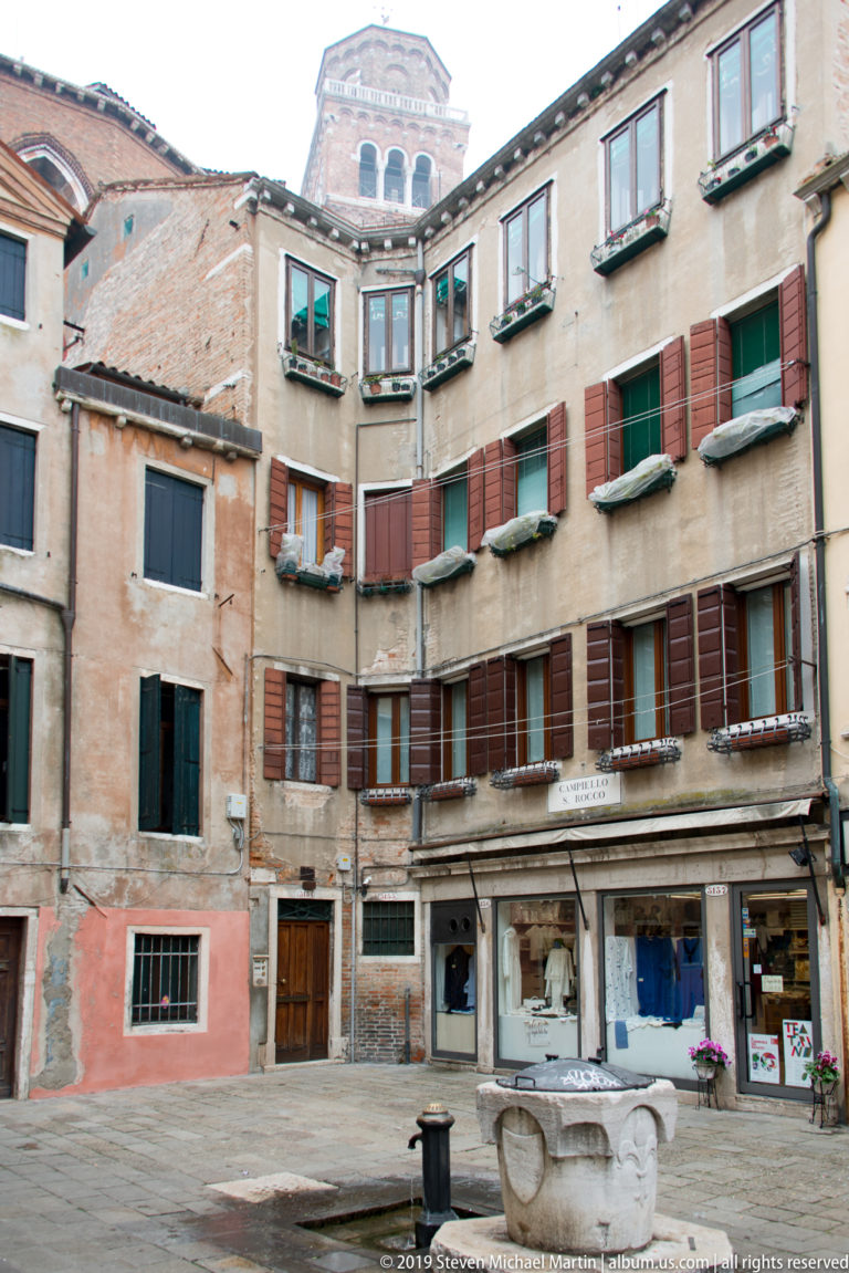 Small Venetian square with a central stone wellhead, weathered multistory housing, shutters, flower boxes, and ground-floor shops.