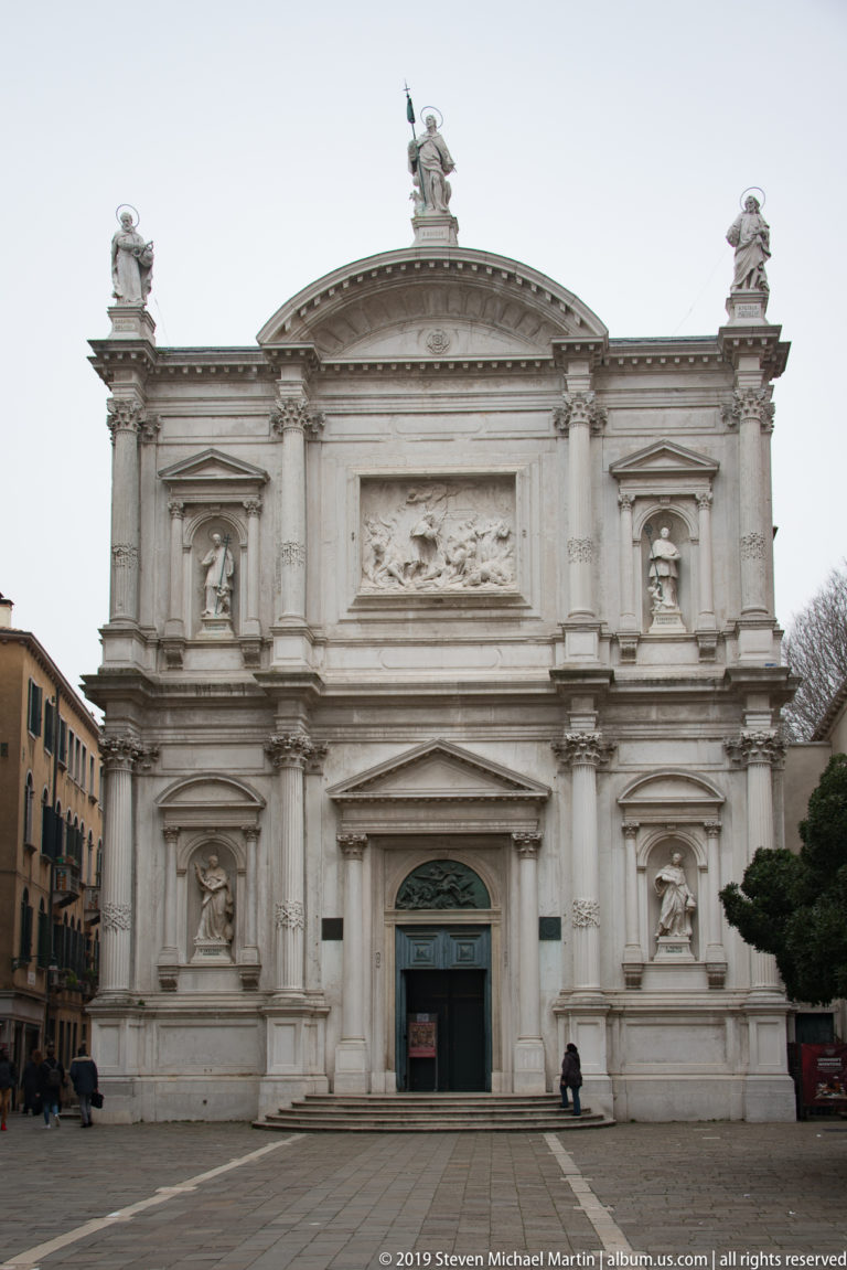 White monumental church façade with central doorway, pediments, sculpted niches, relief panels, and statues facing an open square.