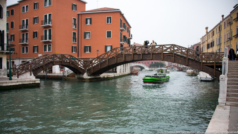 High arched wooden footbridge with pedestrians above a green motorboat passing through a canal between colorful buildings.