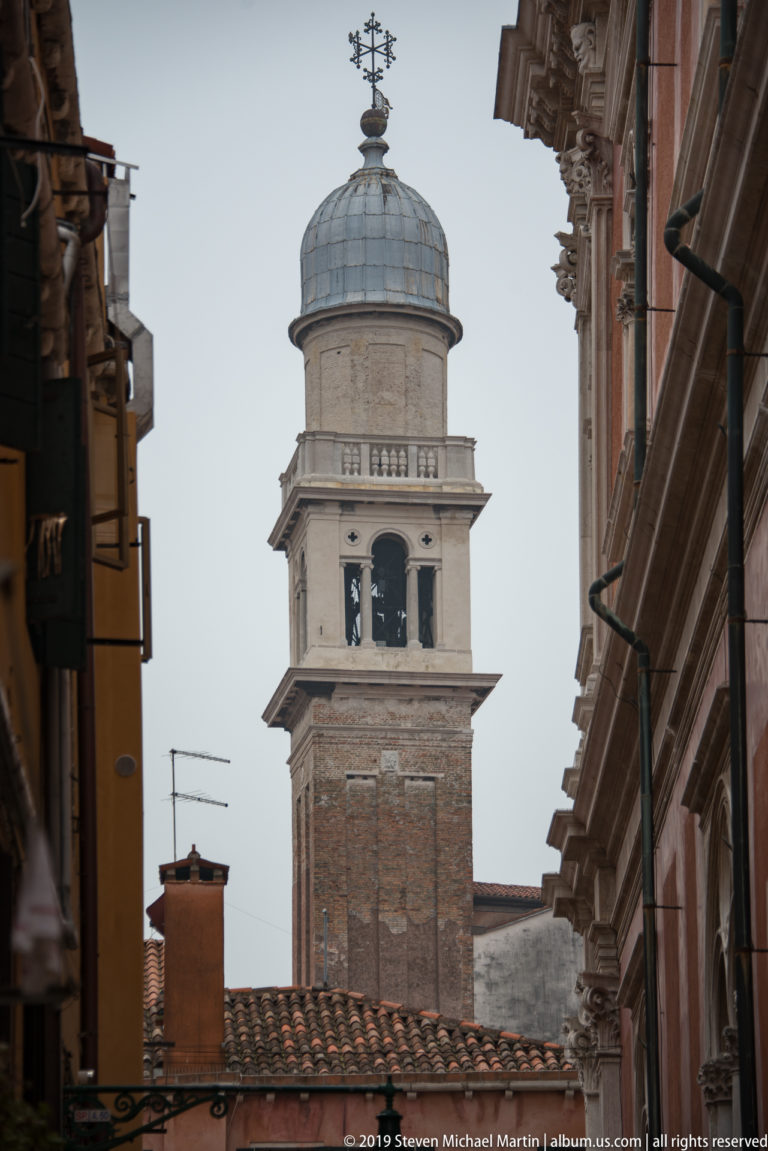 Brick bell tower with an open belfry and pale domed lantern rising above tiled roofs between narrow Venetian buildings.