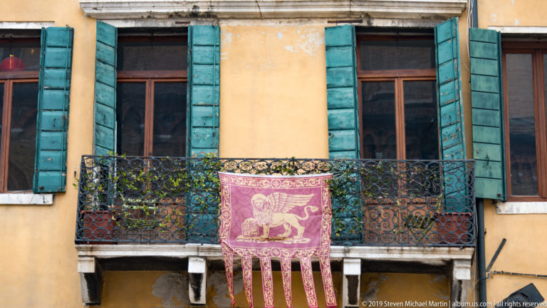Purple winged-lion banner hanging from an iron balcony between turquoise shutters on a yellow Venetian building.