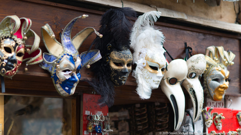 Display of ornate Venetian masks with feathers, metallic decoration, jester forms, and a long white plague-doctor beak.
