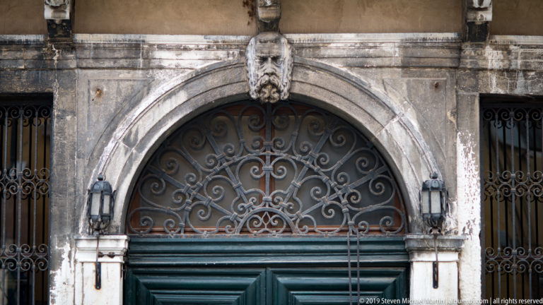 Arched stone doorway with dark green doors, ornate wrought-iron fanlight, carved bearded keystone head, and wall lanterns.