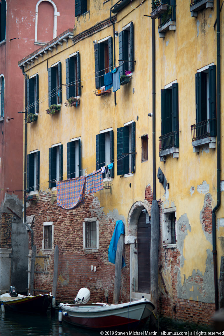 Weathered yellow canal-side building with green shutters, hanging laundry, exposed brick patches, and a small boat below.