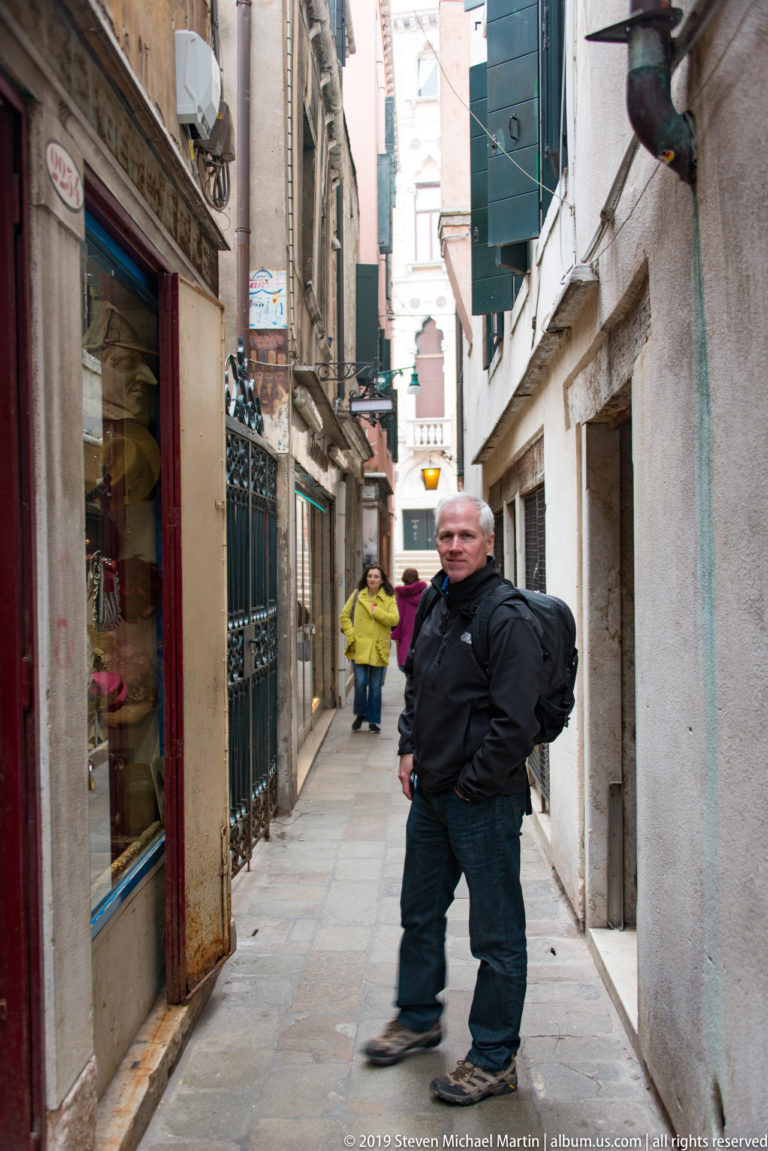 Man walking through a narrow Venetian pedestrian lane with shopfronts, stone walls, shutters, and other people farther ahead.