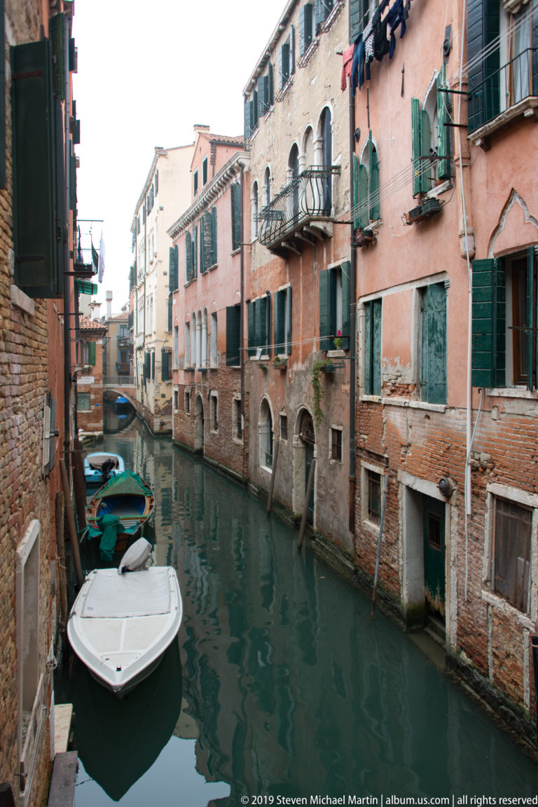 Narrow canal lined by weathered pink and brick residential buildings with green shutters, balconies, laundry, and small boats.