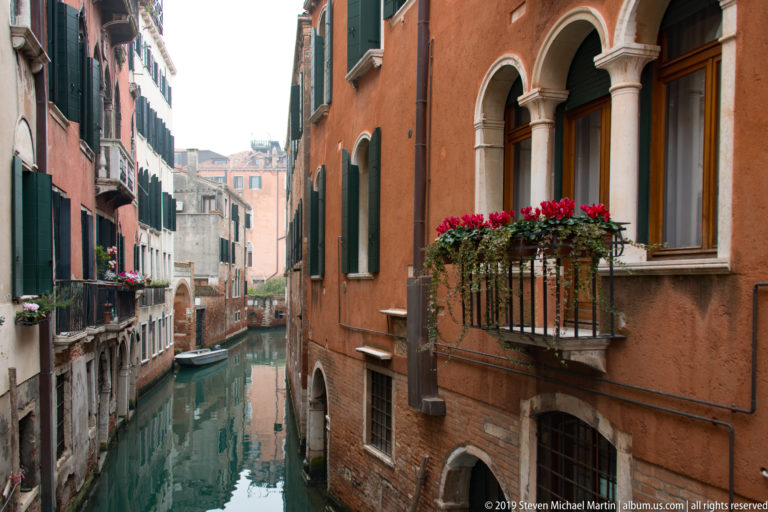Quiet canal beside a terracotta building with arched windows, green shutters, and a balcony filled with red flowers.