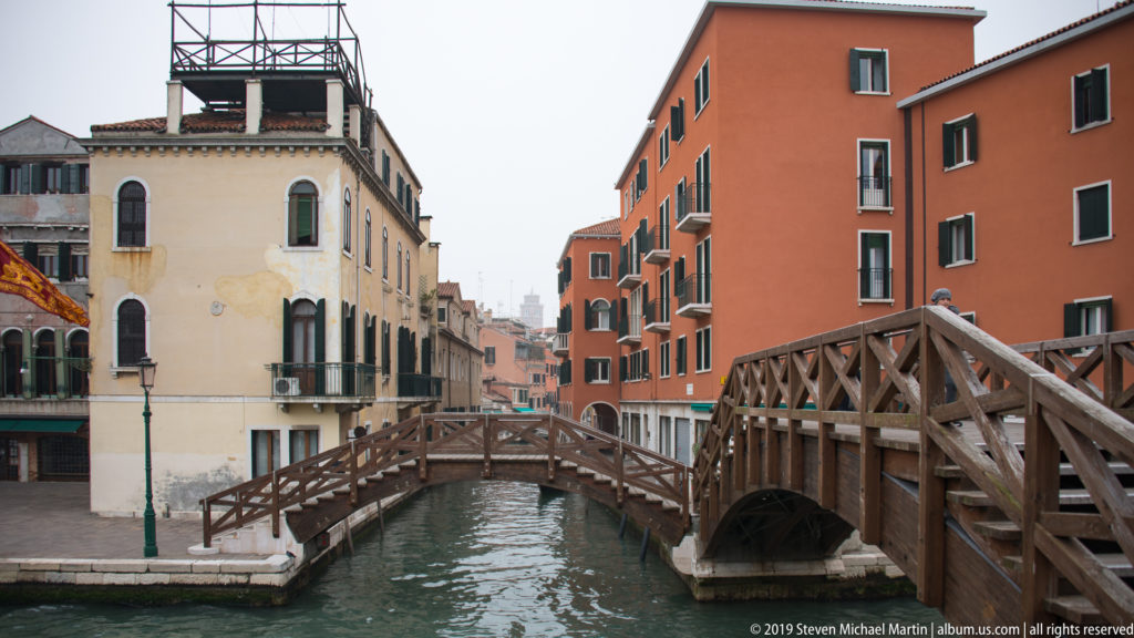 Arched wooden pedestrian bridge with cross-braced railings spanning a narrow canal between cream and terracotta buildings.