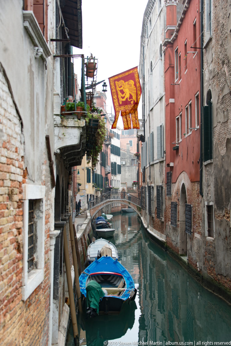Narrow canal with small moored boats beneath a red winged-lion banner between weathered brick and stucco buildings.