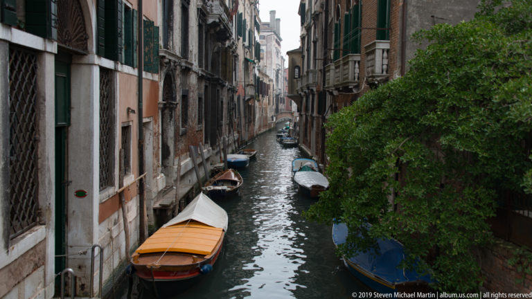 Narrow Venetian canal extending between tall residential buildings, with moored boats and a leafy tree along the water.