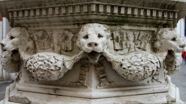 Close view of weathered stone animal heads and carved garlands projecting from the corner of an architectural base.