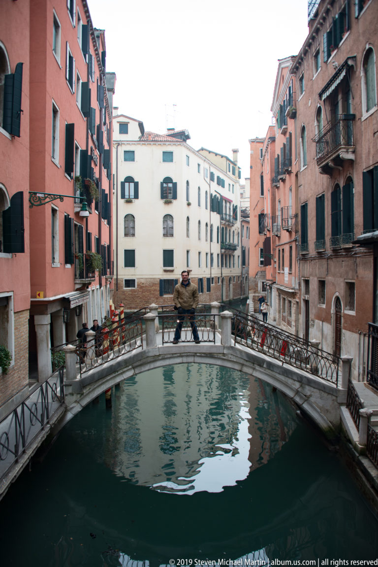 Low arched pedestrian bridge reflected in a quiet canal between red, white, and brick residential façades.