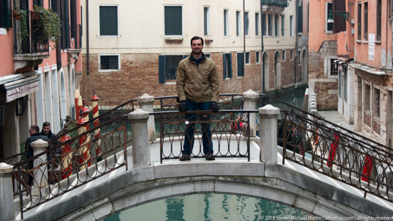 Man standing on a small arched stone footbridge with iron railings above a narrow residential canal.