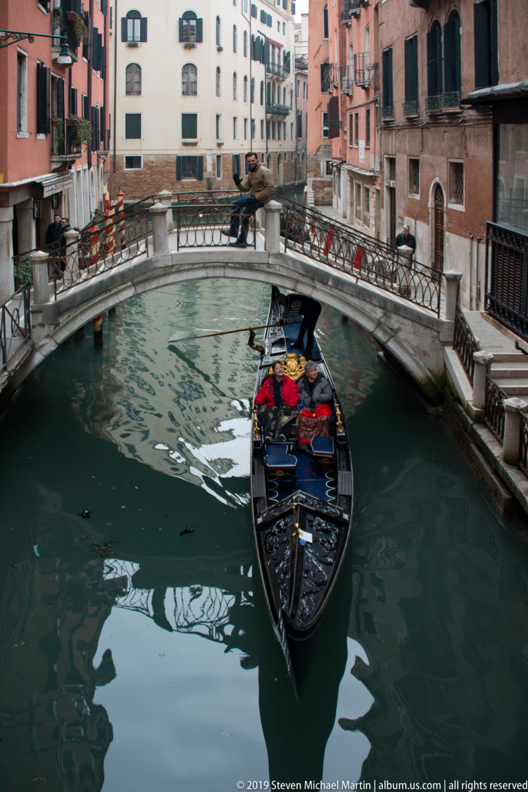 Black gondola with seated passengers approaching a low arched footbridge on a narrow green-water canal.