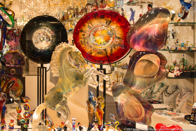 Colorful blown-glass plates, vessels, abstract forms, and small figurines arranged in a brightly lit Venetian shop display.