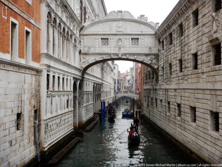 Bridge of Sighs above a narrow canal with gondolas, pale arcades on the left, and heavy stone walls on the right.