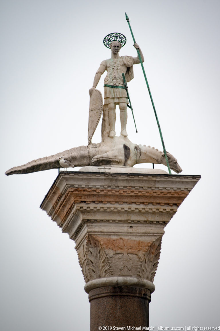Stone figure of Saint Theodore with spear and shield standing above a dragon-like creature atop a monumental column.