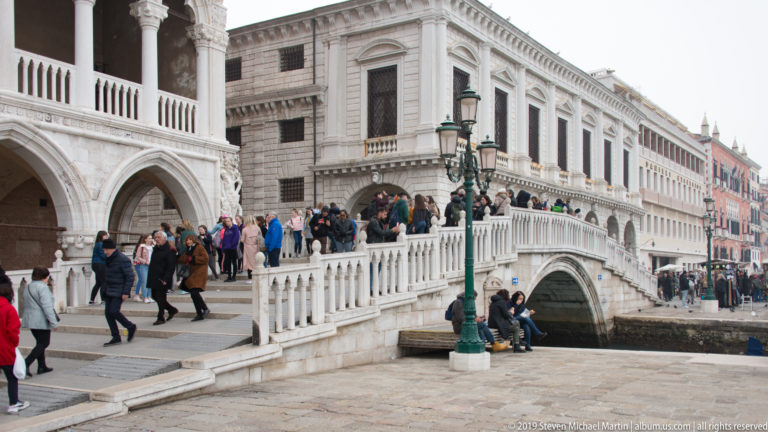 Crowds walking on the stone steps of the Rialto Bridge, with its large canal arch and built upper range visible.