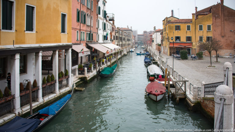 Long narrow canal between colorful Venetian buildings, with covered boats moored along both sides and pedestrians near the quays.