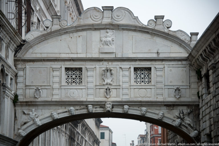 Close view of the Bridge of Sighs showing carved stone panels, pierced windows, shields, and the upper curve of the canal arch.
