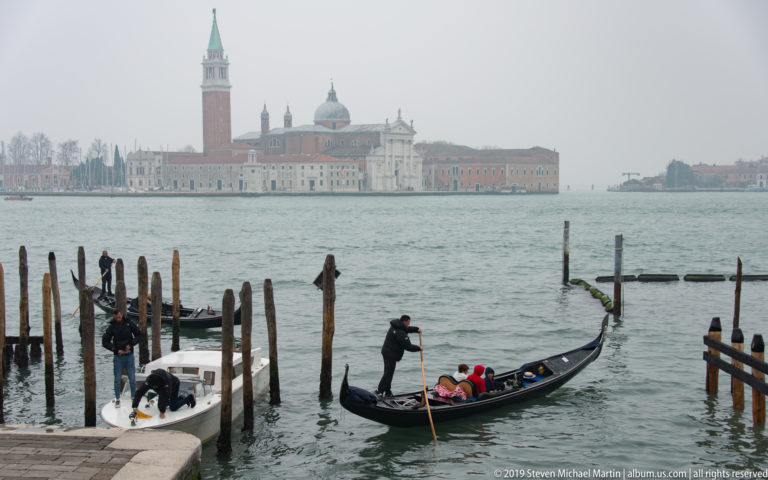Gondolier carrying passengers past wooden mooring posts, with San Giorgio Maggiore and its campanile across the hazy lagoon.