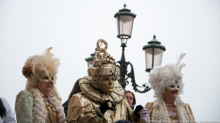 Three Venetian Carnival participants in ornate masks, feathered headpieces, and richly decorated costumes beneath street lamps.