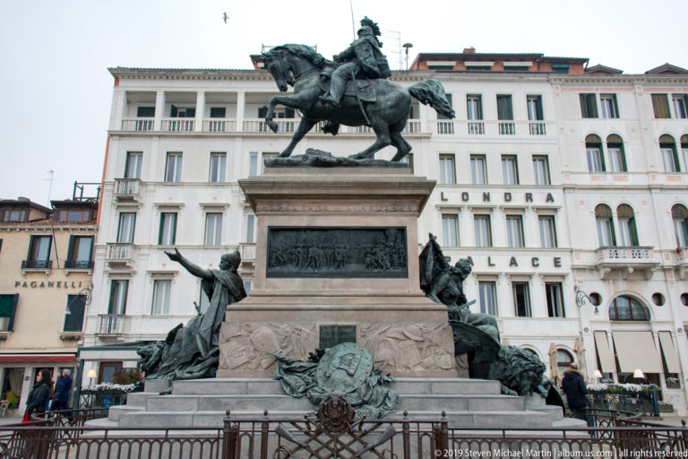 Equestrian monument to Vittorio Emanuele II on a tall stone pedestal, with bronze figures and Venetian buildings behind it.