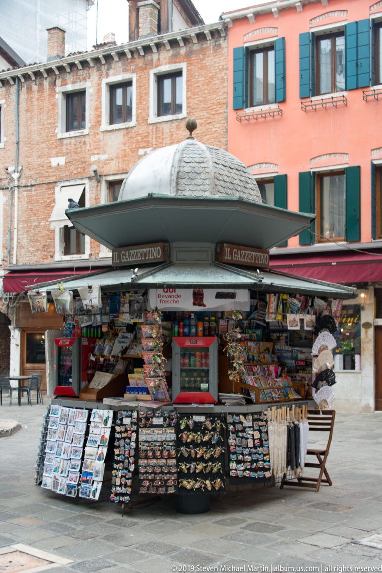 Il Gazzettino newsstand with a domed canopy displays newspapers, postcards, and souvenirs against brick and colorful Venetian buildings.