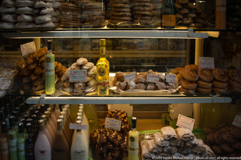 Glass shop case filled with pastries, packaged sweets, bottles of liqueur, and handwritten price cards under warm lighting.