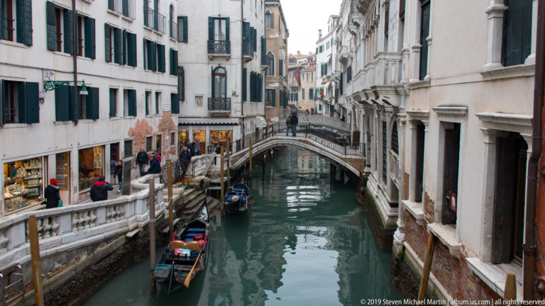 Stone pedestrian bridge over a narrow Venetian canal with several gondolas, pedestrians, shops, and pale historic buildings.