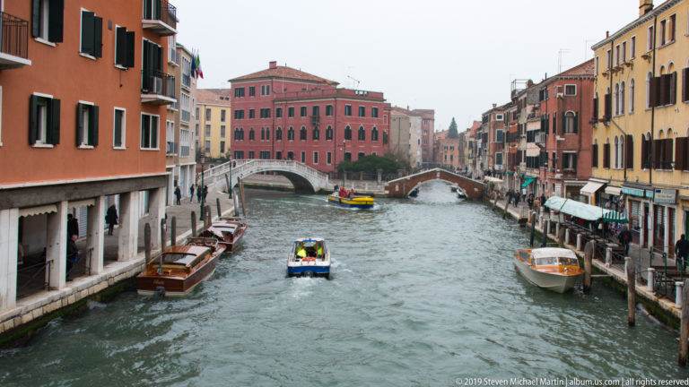 Broad Venetian canal with several motorboats, moored vessels, colorful buildings, and two arched pedestrian bridges in the distance.