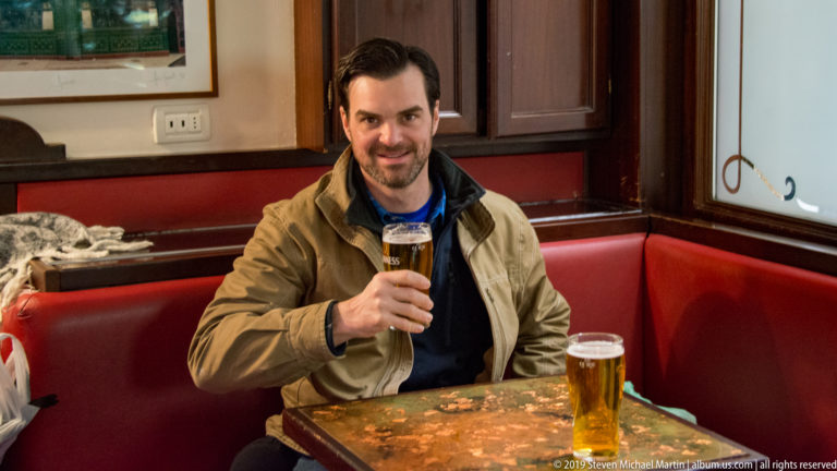 Man seated in a red pub booth holds up a pint of dark beer, with a second golden beer on the table.