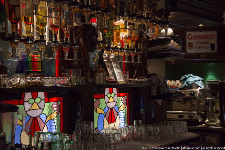 Close view of a pub bar with backlit liquor bottles, hanging glassware, equipment, and two colorful stained-glass panels.
