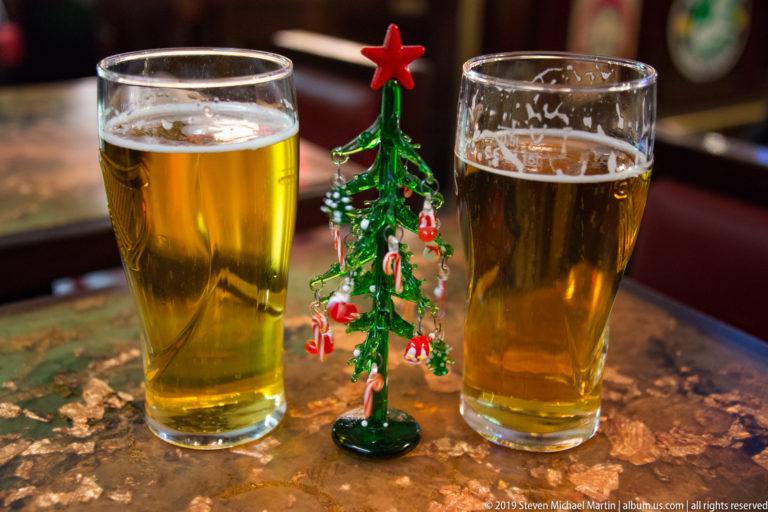 Two glasses of golden beer stand on either side of a small glass Christmas tree topped with a red star.