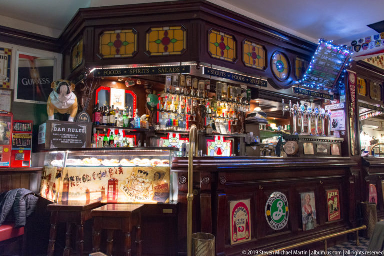 Dark wooden pub bar filled with beer taps, bottles, signs, stained-glass panels, and illuminated displays.