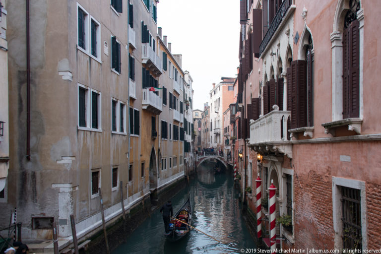 Narrow Venetian canal between tall residential buildings, with a gondola on the water and striped mooring poles at right.
