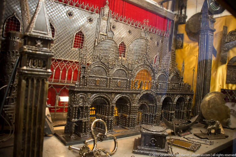 Ornate metal miniature of St. Mark's Basilica displayed against red fabric among other metallic Venice souvenirs.