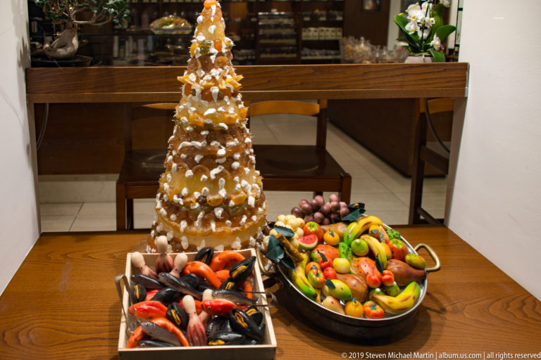 Shop counter with a tall decorated confectionery centerpiece and trays of colorful fruit- and vegetable-shaped sweets.