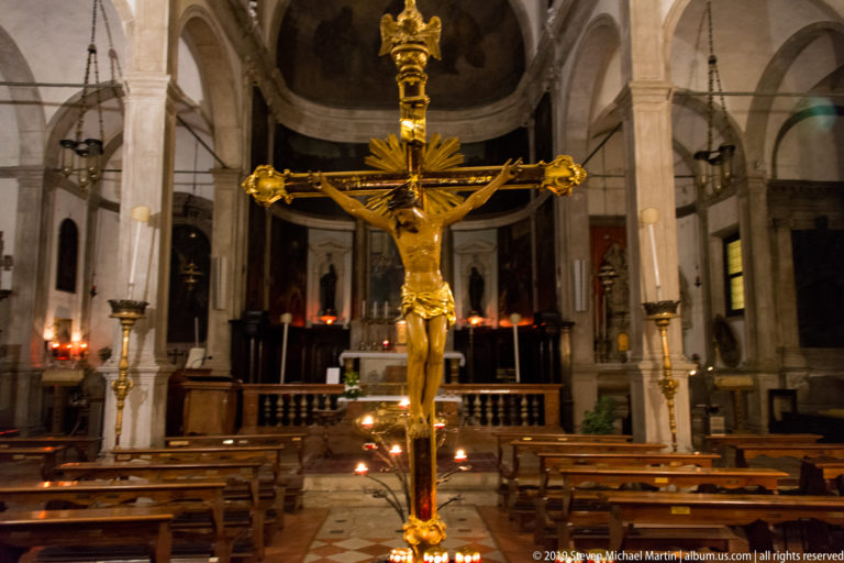 Gilded crucifix centered before a dim church sanctuary, with wooden pews, stone columns, hanging lamps, and votive candles.
