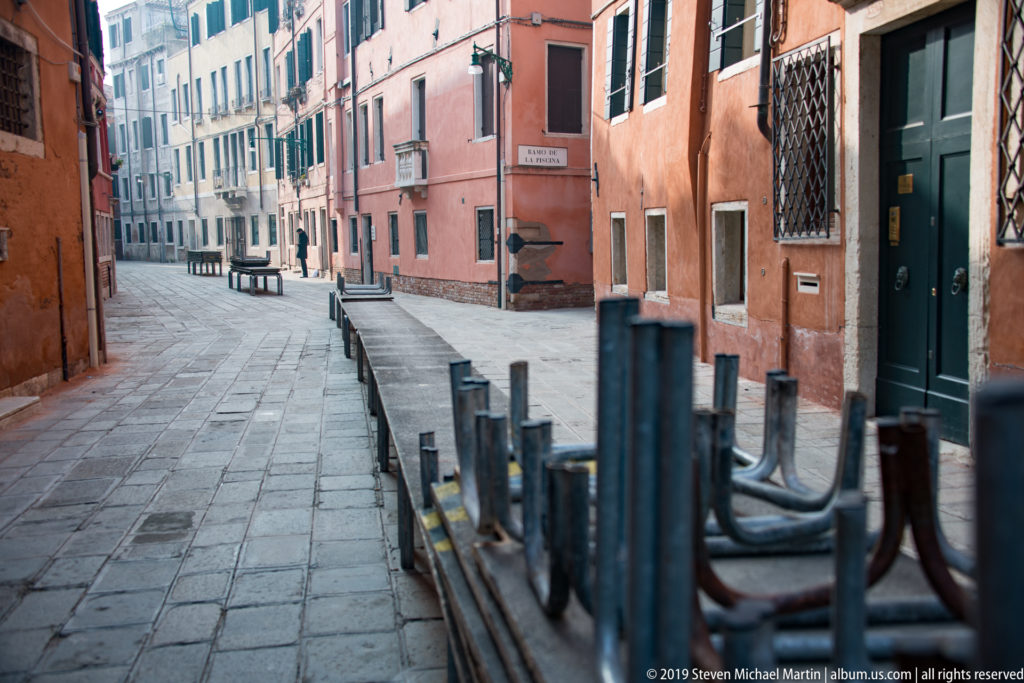 Narrow Venetian lane between stucco buildings, with stacked café furniture along the right side and few pedestrians in the distance.