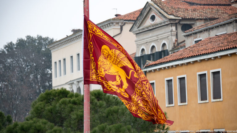 Red and gold Venetian flag with the winged Lion of Saint Mark flying before tiled roofs and city buildings.
